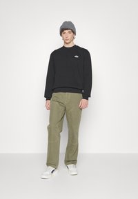 Hanorac negru cu logo mic, pantaloni largi oliv cu detalii de cusătură și adidași albi cu accente bleumarin. Căciulă texturată gri.