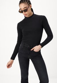 Black long-sleeve top with a ribbed texture and high neck, paired with dark jeans. The model wears tortoiseshell sunglasses and has straight hair.