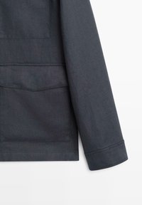 Manche de veste en tissu gris foncé et partie inférieure avec grande poche visible et détail de couture droite au poignet.