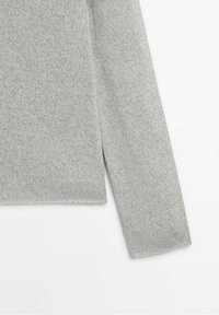 Manche de pull en maille gris clair et détail de la couture latérale sur fond blanc, montrant la texture et le tissage du tissu.