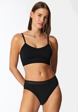 Femme aux cheveux bruns lisses portant un soutien-gorge caraco noir et un sous-vêtement taille haute noir avec une ceinture en dentelle, debout devant un fond blanc.