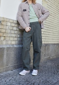 Brun och svart rutigt skjorta över en ljusgrön t-shirt, parat med mörkgröna cargobyxor och ljusrosa sneakers på en kullerstenyta.