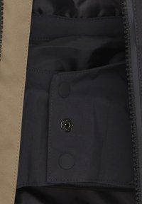 Tessuto esterno beige con una fodera interna scura, dotato di una tasca con bottoni a pressione e dettagli con zip lungo il bordo. Texture liscia in tutto.