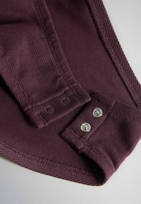 Približni prikaz rebraste ljubičaste tkanine s kopčama na patentni zatvarač na manšeti i području zgloba, prikazujući teksturu i detalje šavova.