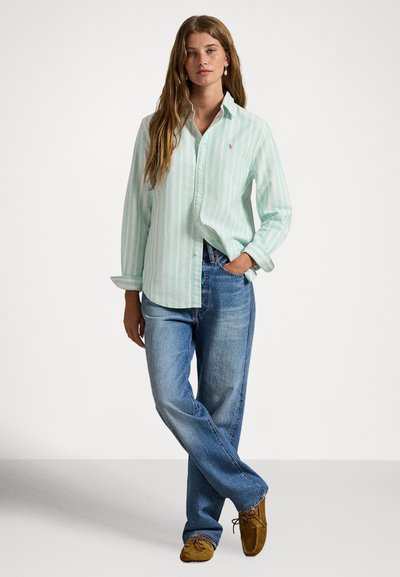 Gaiši zaļa un balta svītraina pogām aiztaisāma krekls ar apkakli, komplektā ar zilām džinsa biksēm un brūnām mokasīnu stila kurpēm.