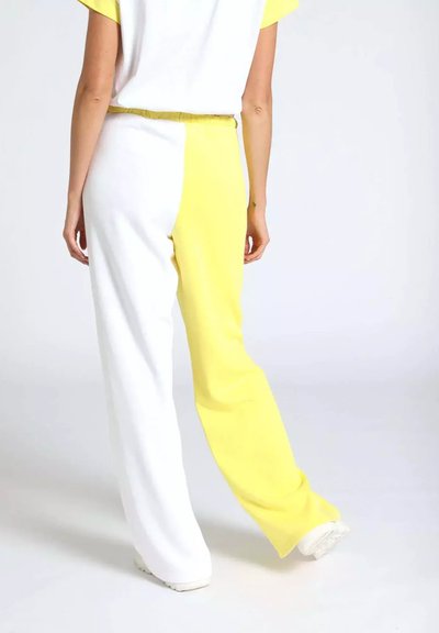 Persona che indossa pantaloni svasati a blocchi di colore bianco e giallo, in piedi davanti a uno sfondo chiaro, vista da dietro.