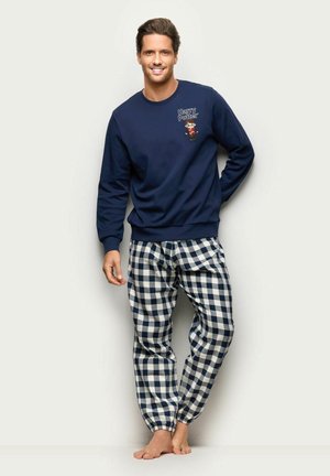 Homme souriant debout, pieds nus, portant un sweat Harry Potter bleu marine et un pantalon de pyjama à carreaux bleu marine et blanc sur fond clair.