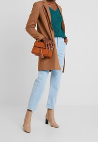 Beżowa marynarka na turkusowym swetrze, jasnoniebieskie dżinsy i cieliste botki za kostkę. Brązowa torebka z detalem w postaci frędzli. Gładkie tekstury i czyste linie.