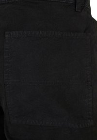 Pantalonii negri au o pungă rectangulară pe spate cu un contur cusut simplu. Materialul pare texturat și rezistent.