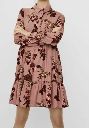 Vrouw draagt een stofroze jurk met lange mouwen, bedrukt met vogel- en bloemenpatronen, staand met gekruiste armen tegen een effen achtergrond.