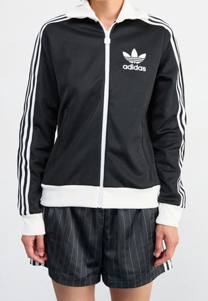 Persoon draagt een zwart Adidas vest met rits met witte strepen op de mouwen en zwarte shorts met fijne strepen.