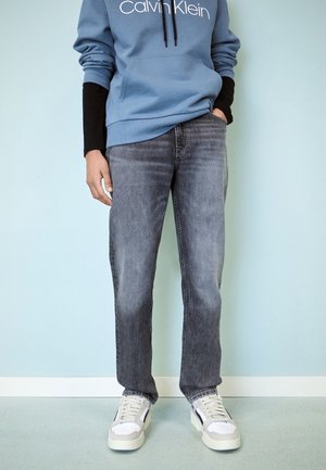 Personne portant un jean noir délavé, des baskets blanches, un sweat à capuche Calvin Klein bleu et un t-shirt à manches longues noir, debout contre un mur bleu clair.