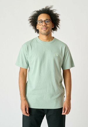Hellgrünes Baumwoll-T-Shirt mit kurzen Ärmeln, Rundhalsausschnitt und einem kleinen gestickten Logo auf der Brust. Wird mit dunklen Hosen getragen.
