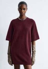 Burgunderfarbenes Strickkleid mit kurzen Ärmeln, geripptem Saum und Rundhalsausschnitt. Kombiniert mit langen, gerafften braunen Lederhandschuhen und einer silbernen Ketten halskette.