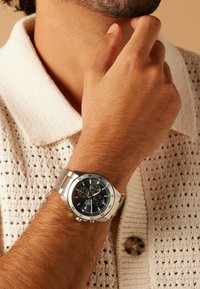 Silver metal wristwatch with a black dial, chronograph features, and contrasting blue accents. Worn on a wrist with light brown skin.