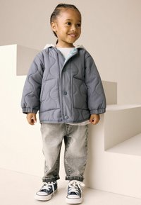 Veste matelassée grise avec une capuche, ornée de boutons bleus et de poches latérales, associée à un jean gris clair et à des baskets bleu marine avec des semelles blanches.