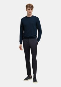 Maglione blu navy a girocollo e pantaloni eleganti scuri. Il maglione presenta maniche texturizzate e un logo sottile. Sneakers grigio scuro con suole chiare.