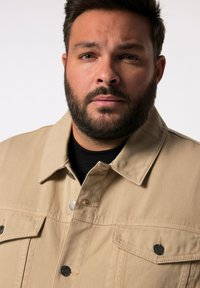 Beige denimová bunda s límcem, dvěma kapsami na hrudi a černými knoflíkovými detaily. Nenošená přes černou košili s texturovaným povrchem.