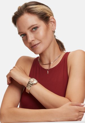 Goldfarbene Armbanduhr mit achteckigem Zifferblatt und Metallarmband, geschichtet mit filigranen Ketten und einer Punktanhängerhalskette auf einem bordeauxfarbenen Oberteil.