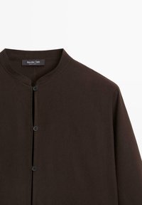 Donkerbruine knoopshirt met een hoge kraag, gemaakt van zacht materiaal. Heeft een eenvoudig ontwerp en minimale stikdetails.