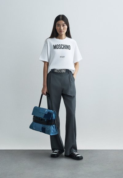 Γυναίκα στέκεται φορώντας ένα λευκό t-shirt Moschino, γκρι παντελόνι με ζώνη Moschino, μαύρα αθλητικά παπούτσια, κρατώντας στην πλευρά της μια μπλε τσάντα patchwork.