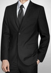 Costume noir avec une fermeture à un seul bouton, revers crantés et deux poches latérales. Chemise blanche et cravate noire complètent le look formel.