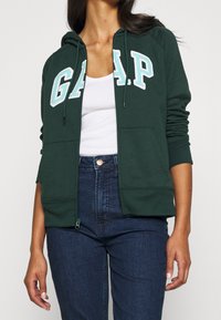 Felpa verde con zip realizzata in tessuto morbido, caratterizzata da un logo "GAP" bianco e azzurro chiaro sulla parte anteriore e tasche frontali.