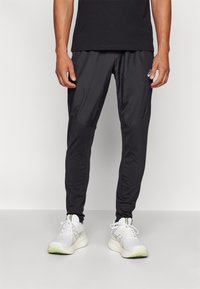 Pantalons de sport noirs à coupe fuselée, en tissu lisse. Comprend des poches latérales et des panneaux élastiques. Portés avec des chaussures de course blanches.