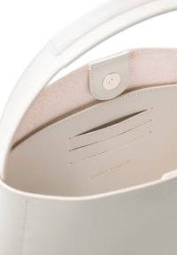 Das Innere der weißen Lederhandtasche hat eine glatte Textur, mit Kartenfächern und einem Druckverschluss. Goldfarbener, geprägter Markenname am Boden.