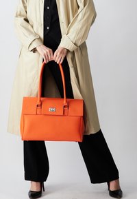 Orange læderhåndtaske med struktureret form, top-håndtag og sølvfarvede hardware-detaljer. Båret med en beige frakke og sort outfit.