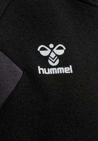 Logo d'abeille stylisé blanc et texte "hummel" imprimés sur un tissu noir avec un détail en tissu gris foncé sur le côté gauche.