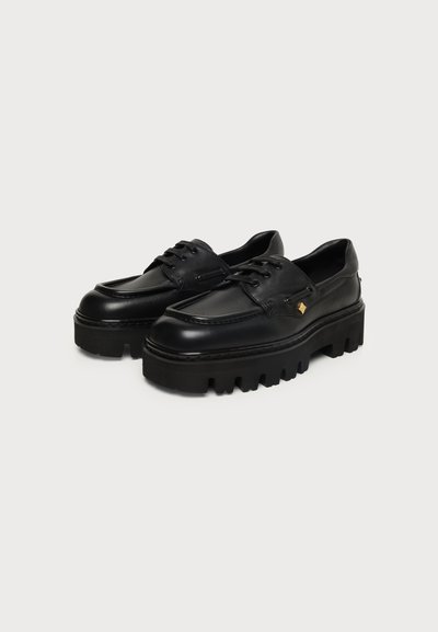 Paire de chaussures en cuir noires épaisses à plateforme avec semelles épaisses crantées et lacets à l'avant sur fond clair uni.