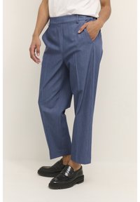Pantalon bleu sur mesure avec un design plissé, fabriqué dans un tissu texturé. Comprend des poches latérales et une coupe droite. Porté avec des mocassins noirs.