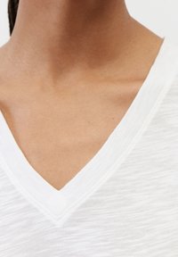 Bílé tričko s výstřihem do V vyrobené z texturované látky. Výstřih má jemný, kontrastní šev. Minimalistický design bez vzorů nebo akcentů.