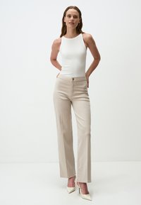 Pantaloni beige a gamba larga con una texture liscia, vita alta, chiusura frontale con bottone e un tessuto leggero. Abbinati a un top aderente bianco.