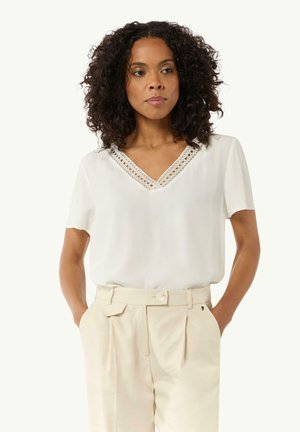 Blouse blanche à manches courtes avec un col en V et des détails en dentelle. Assortie à un pantalon beige taille haute, doté d'un bouton et de plis.