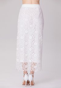 Jupe midi en dentelle blanche avec des motifs floraux, une taille lisse et un ourlet complexe. Portée avec des sandales à talons hauts blanches.