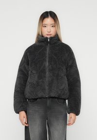Ung kvinde iført en mørkegrå, fuzzy, lynl�åsjakke og slidte sorte jeans, der står op ad en ensfarvet lys baggrund.