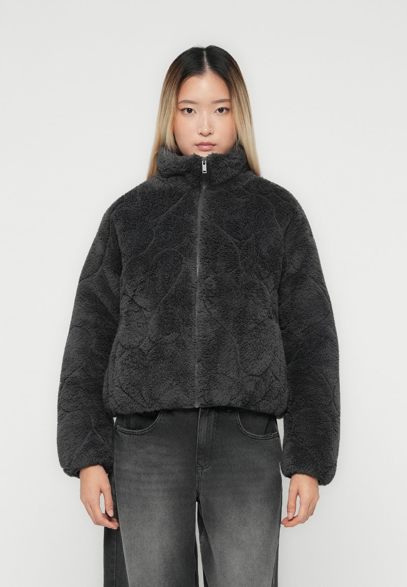 Ung kvinde iført en mørkegrå, fuzzy, lynlåsjakke og slidte sorte jeans, der står op ad en ensfarvet lys baggrund.