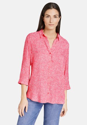 Woman wearing a red button-up shirt with white abstract patterns and three-quarter sleeves, paired with blue jeans against a plain background.