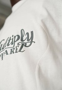 Bílá bavlněná tričko s výrazným šedým potiskem "Multiply" na straně, hladká textura a lehce vypasovaný střih.
