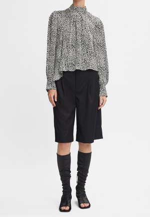 Kvinde iført en hvid og sort polkaprikket bluse med lange ærmer, sorte knælange shorts og sorte åbne høje støvler, står på en hvid baggrund.