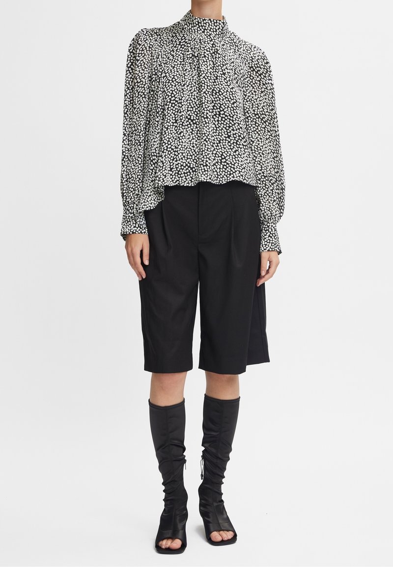 Femme portant un chemisier blanc à pois noirs à manches longues, un short noir au genou et des bottines noires à bout ouvert, debout sur un fond blanc.