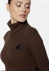 Brązowy sweter z golfem o prążkowanej strukturze, z czarną skórzaną naszywką z logo na lewej piersi. Miękka textura i dopasowany krój.