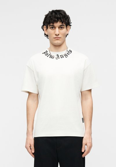 Νέος άνδρας με καστανά σγουρά μαλλιά, φορώντας λευκό μπλουζάκι Palm Angels με μαύρο κείμενο κοντά στον λαιμό και μαύρο παντελόνι, στέκεται μπροστά σε λευκό φόντο.