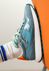 Zapatilla retro con parte superior en color azul verdoso, acentos de ante y brillantes, cordones en menta y detalle del talón en naranja. Suela blanca con tracción verde.