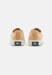 Beežid kanvas sneakersid madala disainiga, valge kummist tallaga ja roheline Lacoste logo kandadel. Sile tekstuur, minimaalne kaunistus.