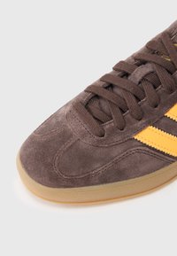 Smeđa cipelica od antilopa sa žutim prugama, s teksturiranim gornjim dijelom, okruglim vrhom i gumennim potplatom. Dizajn s vezicama i ojačanim šavovima.