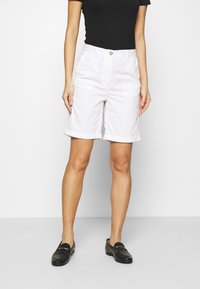Pantaloni corti bianchi in cotone con orlo arrotolato, dotati di chiusura frontale con bottone e zip. Abbinati a mocassini neri e un top nero aderente.