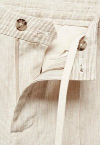 Gros plan de la ceinture d’un pantalon beige rayé avec bouton, boutonnière, fermeture éclair et lien blanc.